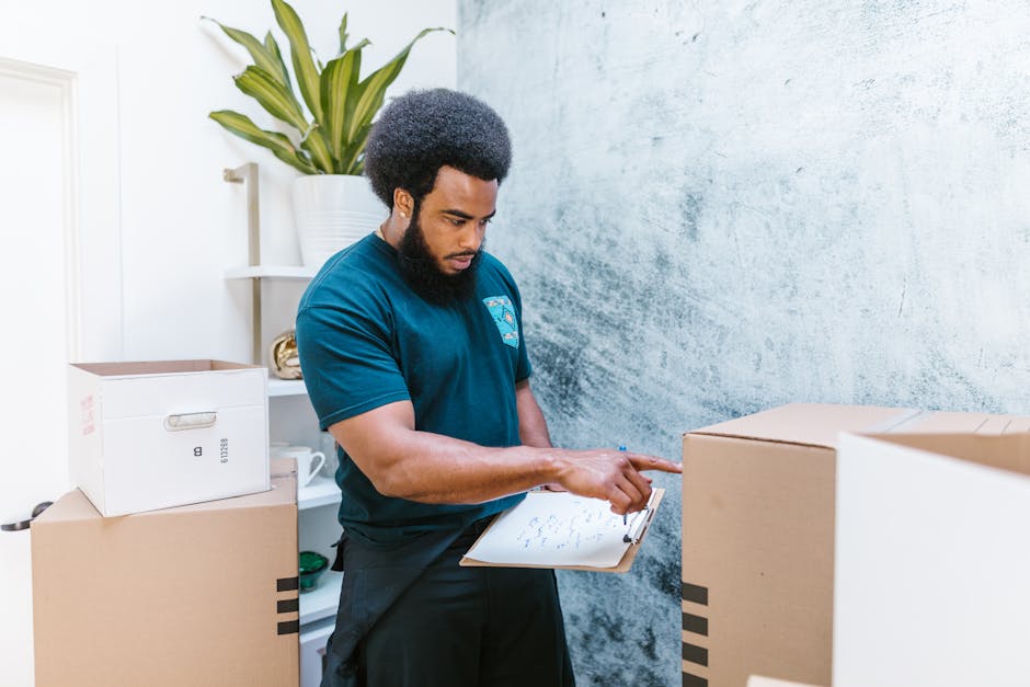 A person wearing a white t-shirt and blue jeans is kneeling on the floor during a home relocation process, writing in red marker on top of a closed cardboard box secured with white packing tape. The i